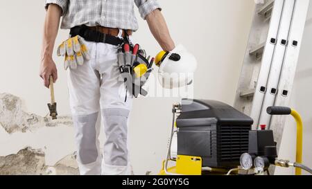 construction man worker wearing a tool bag belt, helmet with headphones and hammer in his hands, gloves in your pocket, compressor  and ladder on whit Stock Photo