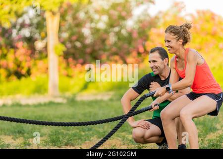 Smiley woman using a battle rope in the park and working out with her personal trainer. Stock Photo