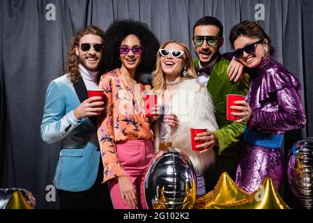 smiling interracial friends in sunglasses standing with plastic cups on grey background Stock Photo