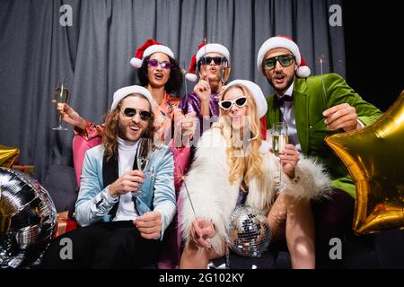 interracial friends in santa hats celebrating new year near grey curtain on black background Stock Photo