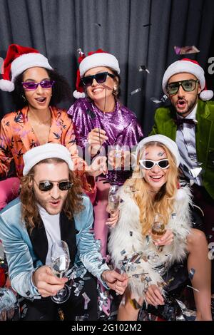 young adult interracial friends in santa hats and sunglasses having party on grey background Stock Photo