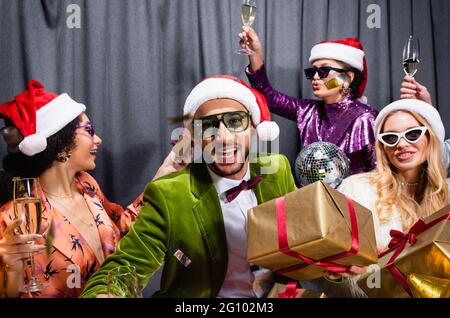smiling interracial friends in santa hats drinking champagne and celebrating new year on grey background Stock Photo