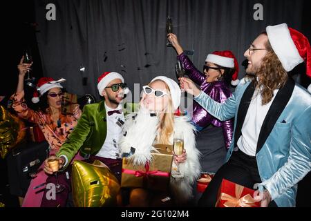 playful interracial friends in santa hats drinking champagne and celebrating new year on grey background Stock Photo