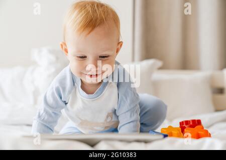 Happy blonde baby boy looking at tablet computer while playing on bed at home Stock Photo