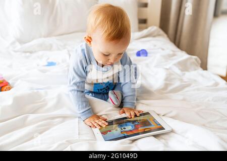 Happy blonde baby boy looking at tablet computer and watching cartoon at home Stock Photo