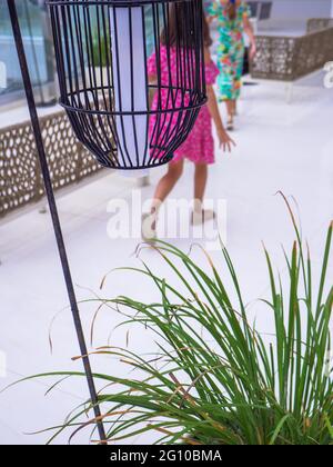 Two girl kids in bright summer dresses running and playing at an open terrace. High black metal grid lantern hanging at the front. Plant in flowerpot. Stock Photo