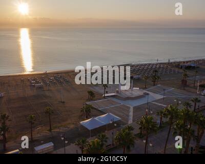 Top aerial view overlooking the sun rising over the calm mediterranean sea with sun path on water surface. Sunbeds with umbrellas, volleyball court. Stock Photo