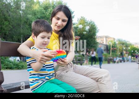 Mom and baby boy 4 years old play with a colored trendy toy Pop it in the park in nature. Antistress sensitive toy or reusable bubble wrap. Trend 2021 Stock Photo