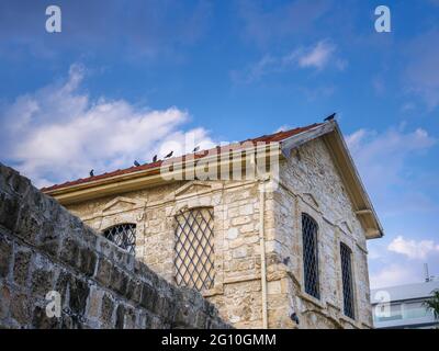 View over the top part of an ancient stone building with red-tiled roof with pigeons against the blue sky and white clouds in Larnaca, Cyprus. Stock Photo