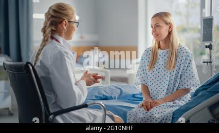 Hospital Ward: Friendly Female Doctor Asks Health Care Questions of Young Woman Patient Sitting on a Bed. Physician Does Checkup, Talks with Happy Stock Photo