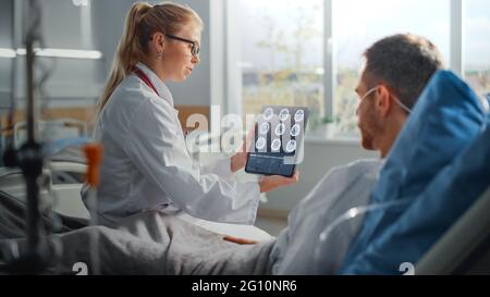 Hospital Ward: Friendly Female Doctor Talks to Sick Male Patient Resting in Bed, Uses Tablet Computer, Shows and Explains Brain CT MRI Scan Results Stock Photo