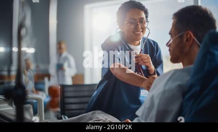 Hospital Ward: Friendly Black Head Nurse Uses Stethoscope to Listen to Heartbeat and Lungs of Recovering Male Patient Resting in Bed, Does Checkup Stock Photo