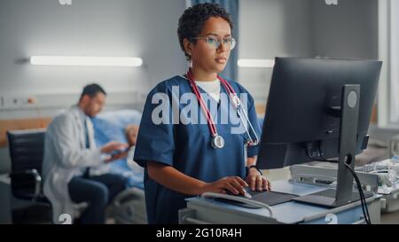 Hospital Ward: Professional Smiling Black Female Head Nurse or Doctor Wearing Stethoscope Uses Medical Computer. In the Background Patients in Beds Stock Photo