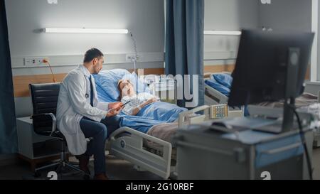 Hospital Ward: in Modern Equipment Clinic Surgeon Using Tablet Computer Consulting Patient Recovering After Successful Surgery in Bed. Modern Advanced Stock Photo