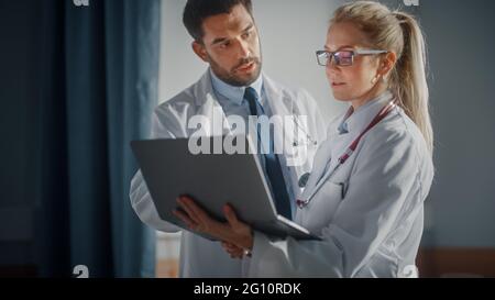 Hospital Ward: Male and Feamle Professional Doctors Hold Laptop Computer, Talk About Patient Medical History, Discuss Health Care Advice, Treatment Stock Photo