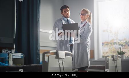 Hospital Ward: Male and Feamle Professional Doctors Hold Laptop Computer, Talk About Patient Medical History, Discuss Health Care Advice, Treatment Stock Photo