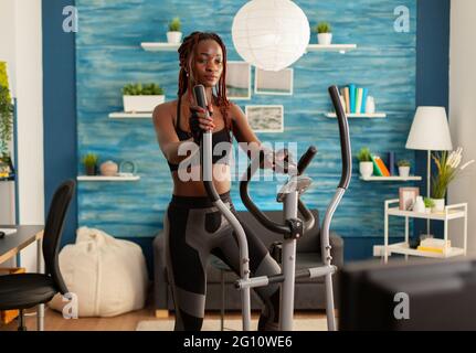 Strong athletic fit black woman doing cross training exercising in home living room using modern eliptic cardio equipment. exercising and changing tv channels. Stock Photo