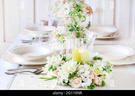 Floral decorations on summer party table. Candle in glass vase, flower wreath around it. White plates, table cloth, romantic wedding reception. Stock Photo