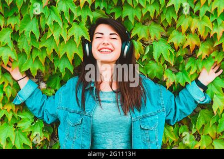 Young hipster girl listens and enjoys music leaning against an ivy wall - Pretty millennial woman relaxes with headphones in a city park Stock Photo