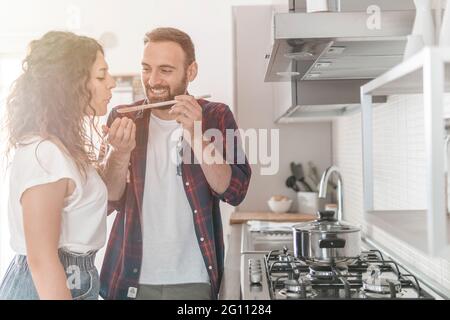 Beautiful young couple smiles while tasting the prepared food - couple has fun in their new kitchen preparing dishes Stock Photo