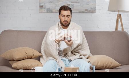 sick man sitting on sofa and holding bottle with pills near coffee table with drinks Stock Photo