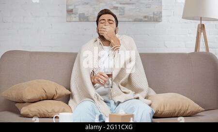 sick man sitting on sofa and taking pill near coffee table with cup and bottles Stock Photo