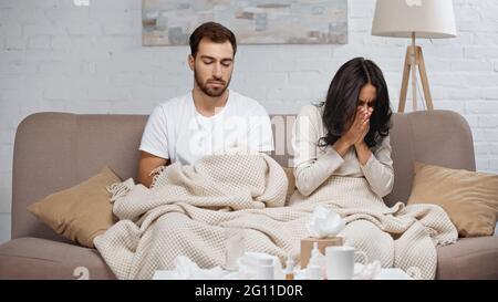 sick woman sneezing near man and bottles with pills on coffee table Stock Photo