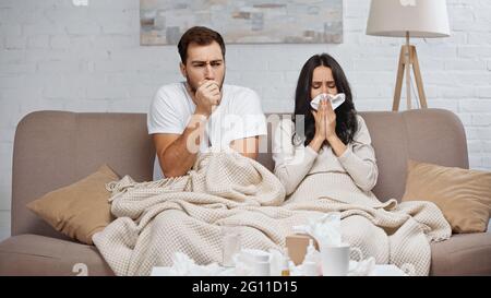 sick woman sneezing while man couching near bottles with pills on coffee table Stock Photo