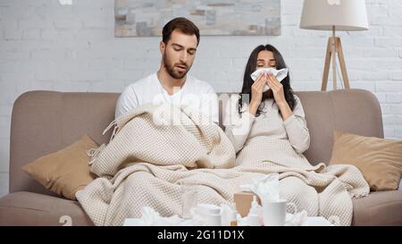 sick woman sneezing in tissue near man and bottles with pills on coffee table Stock Photo