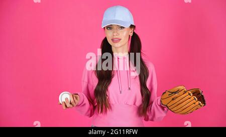 young adult woman in cap and hoodie holding baseball equipment isolated on pink Stock Photo