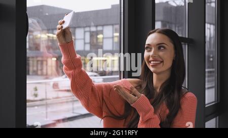 smiling young adult woman taking selfie on cellphone and gesturing with hand near window in modern office Stock Photo