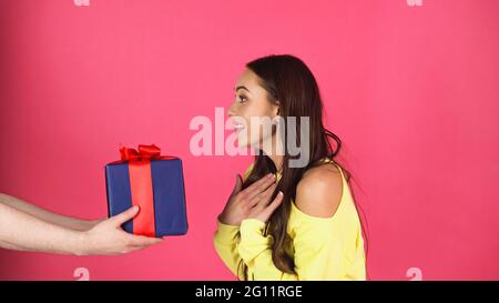 amazed young adult woman standing near male hands with gift box isolated on pink Stock Photo