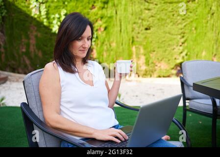A Caucasian brunette woman holds a cup of coffee while working on her laptop Stock Photo