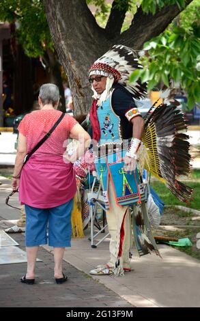 A Native American man dressed in Plains Indian regalia sells artworks and talks with tourists in the historic Plaza in Santa Fe, New Mexico. Stock Photo