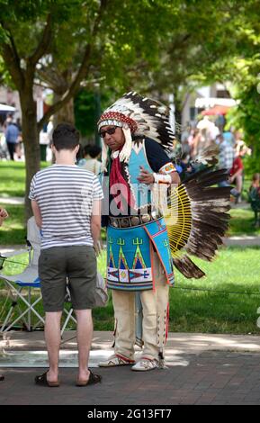 A Native American man dressed in Plains Indian regalia sells artworks and talks with tourists in the historic Plaza in Santa Fe, New Mexico. Stock Photo