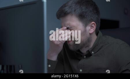 Home office. Handsome man working late at home. Tired young male feels pain, eye strain, dry irritated eyes, fatigue from working at monitor Stock Photo