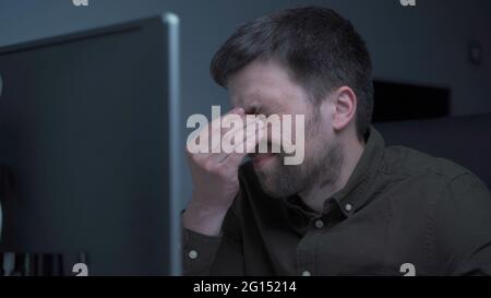 Home office. Handsome man working late at home. Tired young male feels pain, eye strain, dry irritated eyes, fatigue from working at monitor Stock Photo