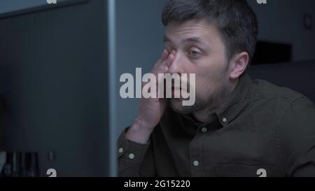 Tired male working on computer. Sad exhausted man at home office. Stressful work, stress at workplace. Software developer looks very tired during Stock Photo