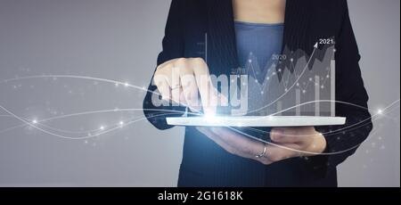 Digital business and stock market. White tablet in businesswoman hand with digital hologram financial charts showing growing revenue In 2021 on grey. Stock Photo