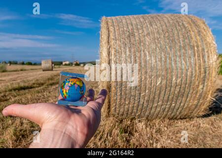 A hand holds a small globe with a hay bales field and the blue sky in the background Stock Photo