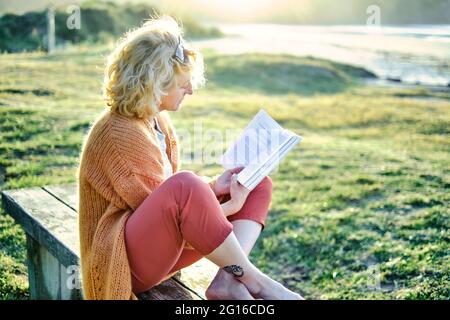 Young mature blonde caucasian woman reading outdoor in a bench in a green landscape close to a beach in a sunny day. Stock Photo