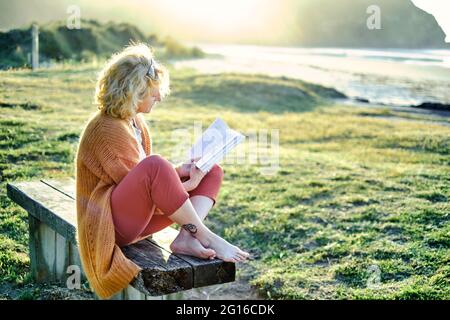 Young mature blonde caucasian woman reading outdoor in a bench in a green landscape close to a beach in a sunny day. Stock Photo