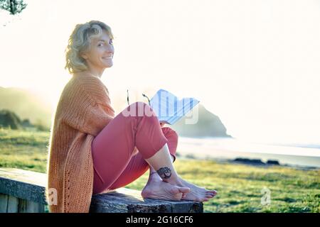 Young mature blonde caucasian woman reading outdoor in a bench in a green landscape close to a beach in a sunny day. Stock Photo