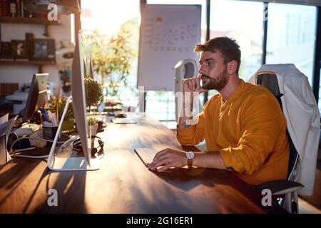 A young male office worker is thinking about job while sitting at the desk in a working atmosphere in the office. Business, office, job Stock Photo