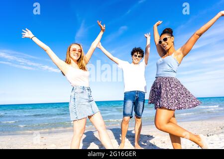 Three very happy friends having fun at the beach posing laughing looking at camera for a portrait with raised arms. Gen z multiracial carefree people Stock Photo