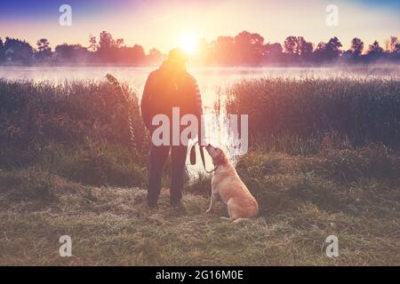 A man with a labrador retriever dog walks in the countryside by the lake on an early autumn morning Stock Photo