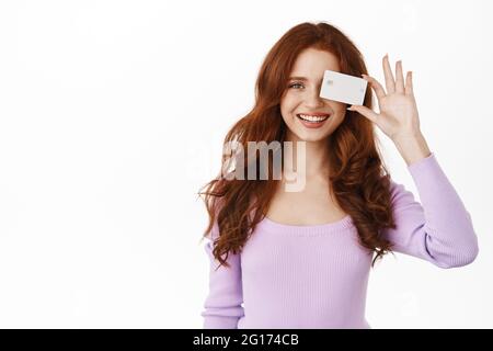 Portrait of beautiful redhead woman smiling white teeth, showing bank credit card, contactless payment, standing against white background Stock Photo