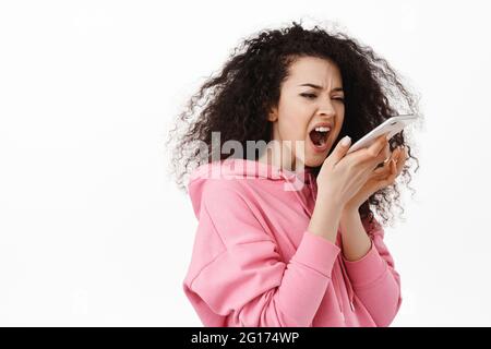Portrait of annoyed and angry woman yelling, screaming at mobile phone, shouting in smartphone speakerphone with furious face, standing against white Stock Photo