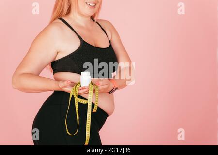 Happy young woman with oversized body wearing yoga outfit holding jar of pills and measuring tape standing on pink background with copy space. Stock Photo