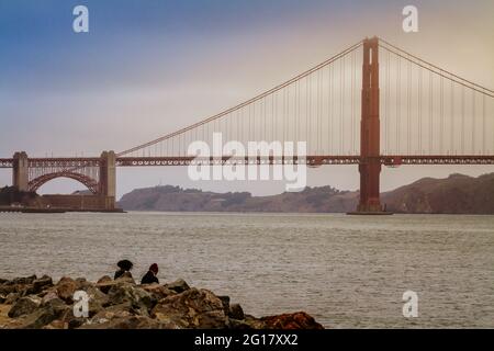 Golden Gate Bridge on a foggy day and two people sitting by San Francisco Bay Stock Photo